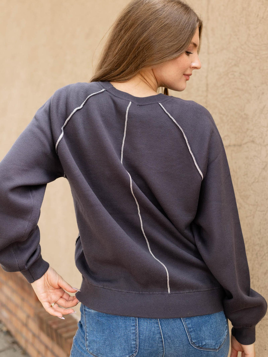A woman wearing a dark charcoal gray crewneck sweatshirt with contrasting white diagonal stitching details, paired with blue jeans, photographed from behind against a textured beige wall.