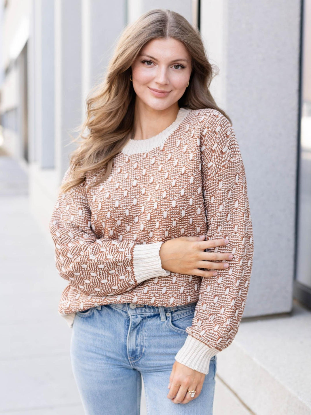 A woman wearing a textured knit sweater with a brown and beige geometric pattern and cream ribbed cuffs, paired with light blue jeans, standing outdoors against a modern gray building.