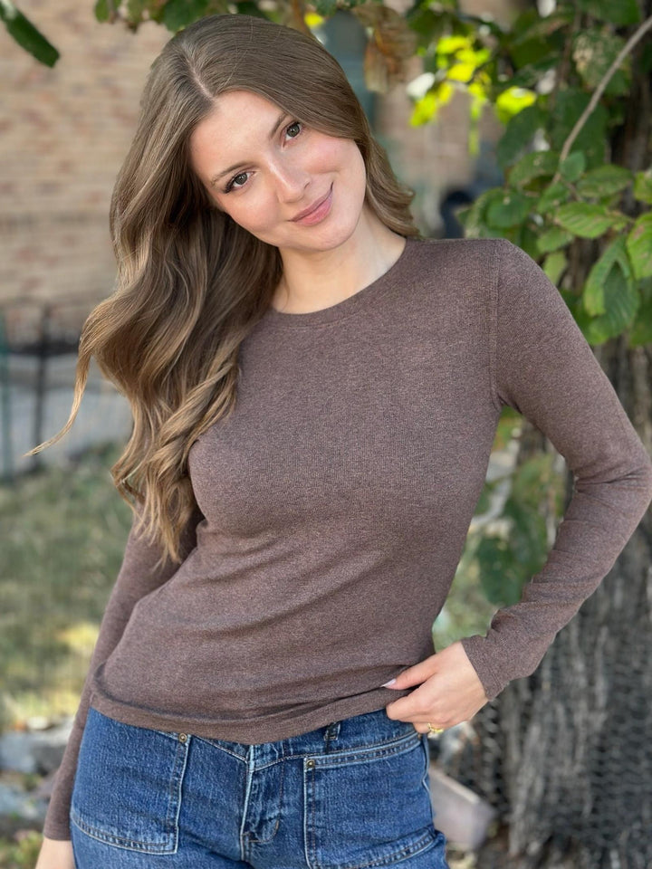 A woman wearing a fitted, long-sleeve crewneck knit top in a heathered taupe-brown color, paired with blue jeans, standing outdoors in front of a brick wall and green foliage.