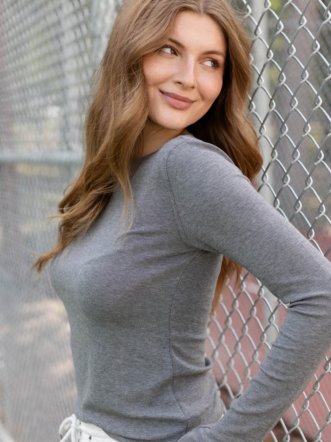 A woman with long light brown hair wearing a fitted heather gray long-sleeve top, posing in front of a chain-link fence.