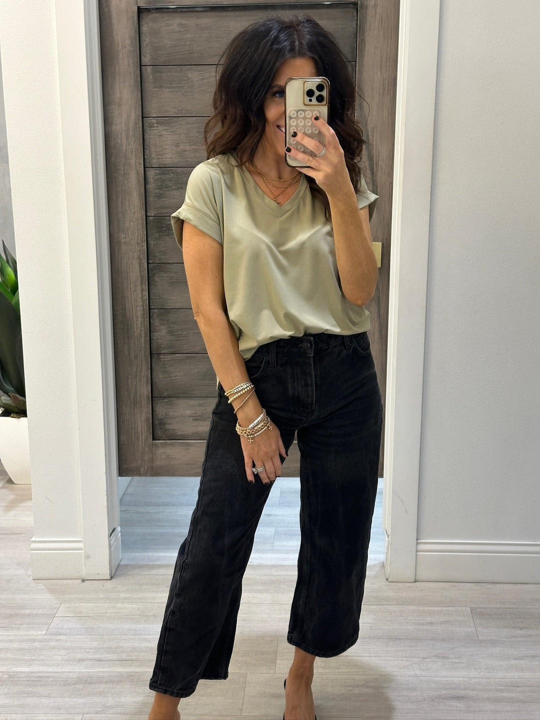 Woman taking a mirror selfie wearing a green top and black pants in a home setting.