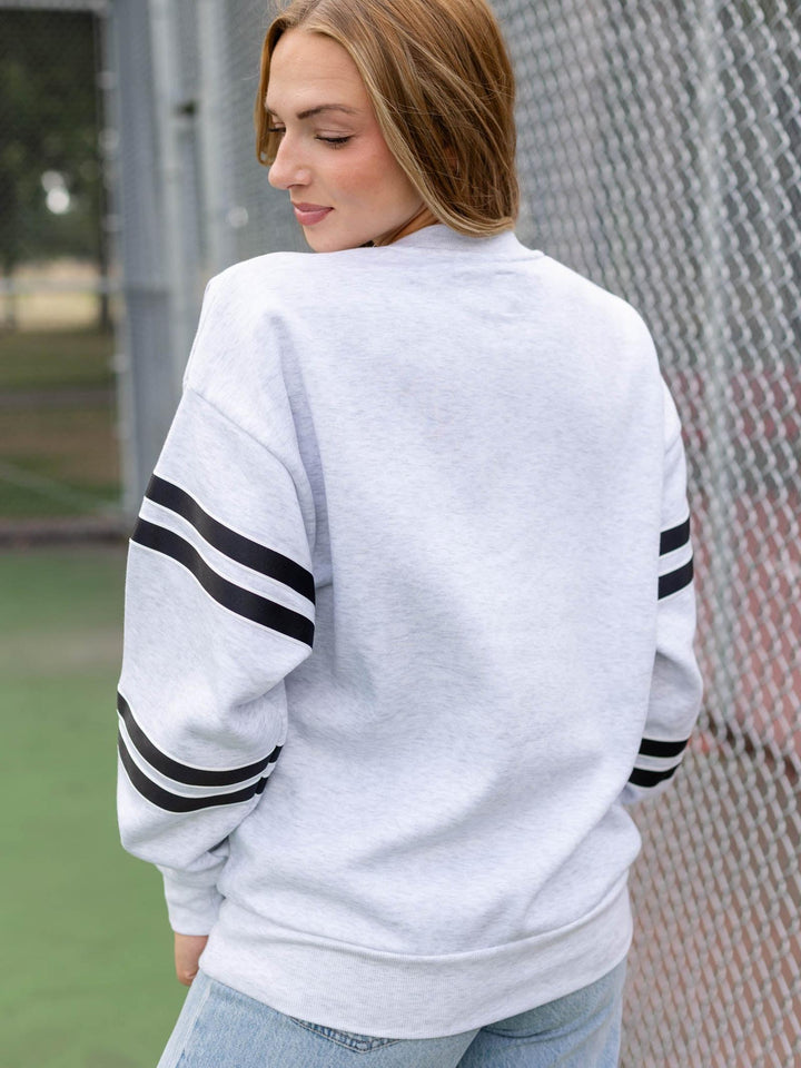 A woman wearing a light gray heather crewneck sweatshirt with black athletic stripes on the sleeves, photographed from the back in an outdoor setting with a chain-link fence.