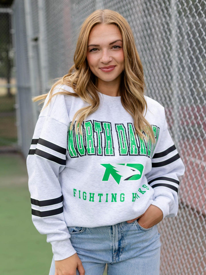 A woman wearing a white North Dakota Fighting Hawks sweatshirt with green and black lettering and sleeve stripes, paired with light blue jeans, standing in front of a chain-link fence.