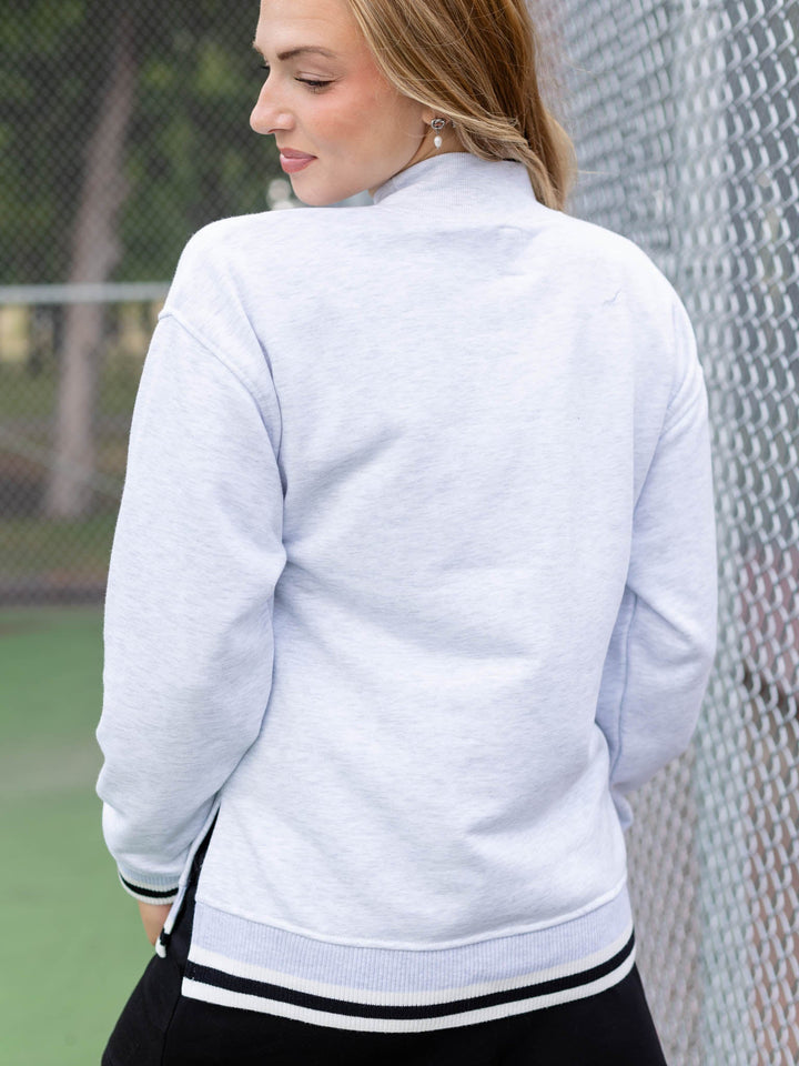 A woman viewed from the back wearing a light heather gray mock-neck athletic sweatshirt with black and white striped ribbed trim, standing outdoors near a chain-link fence.