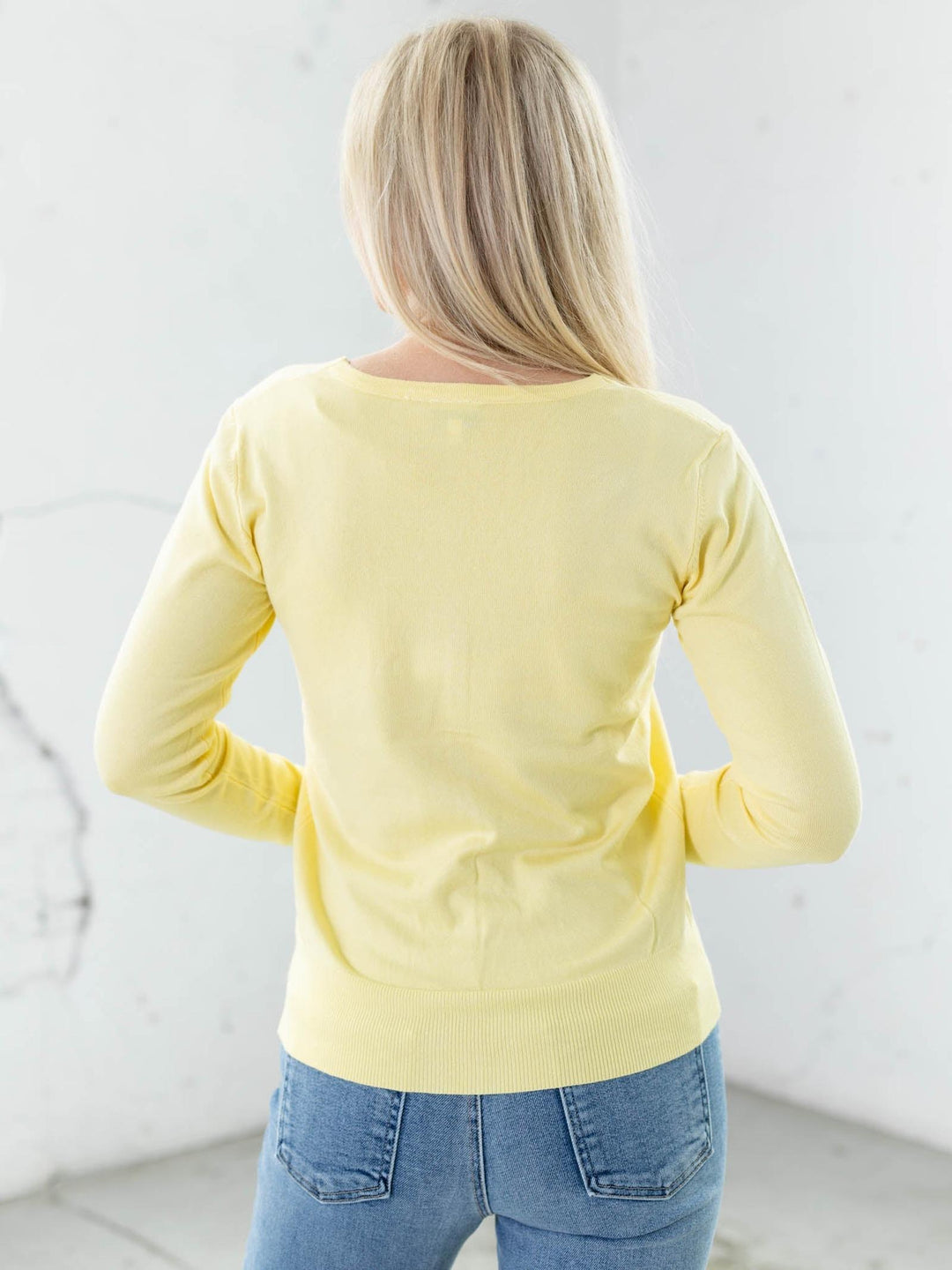 Person wearing a yellow long-sleeve top and blue jeans against a white wall.