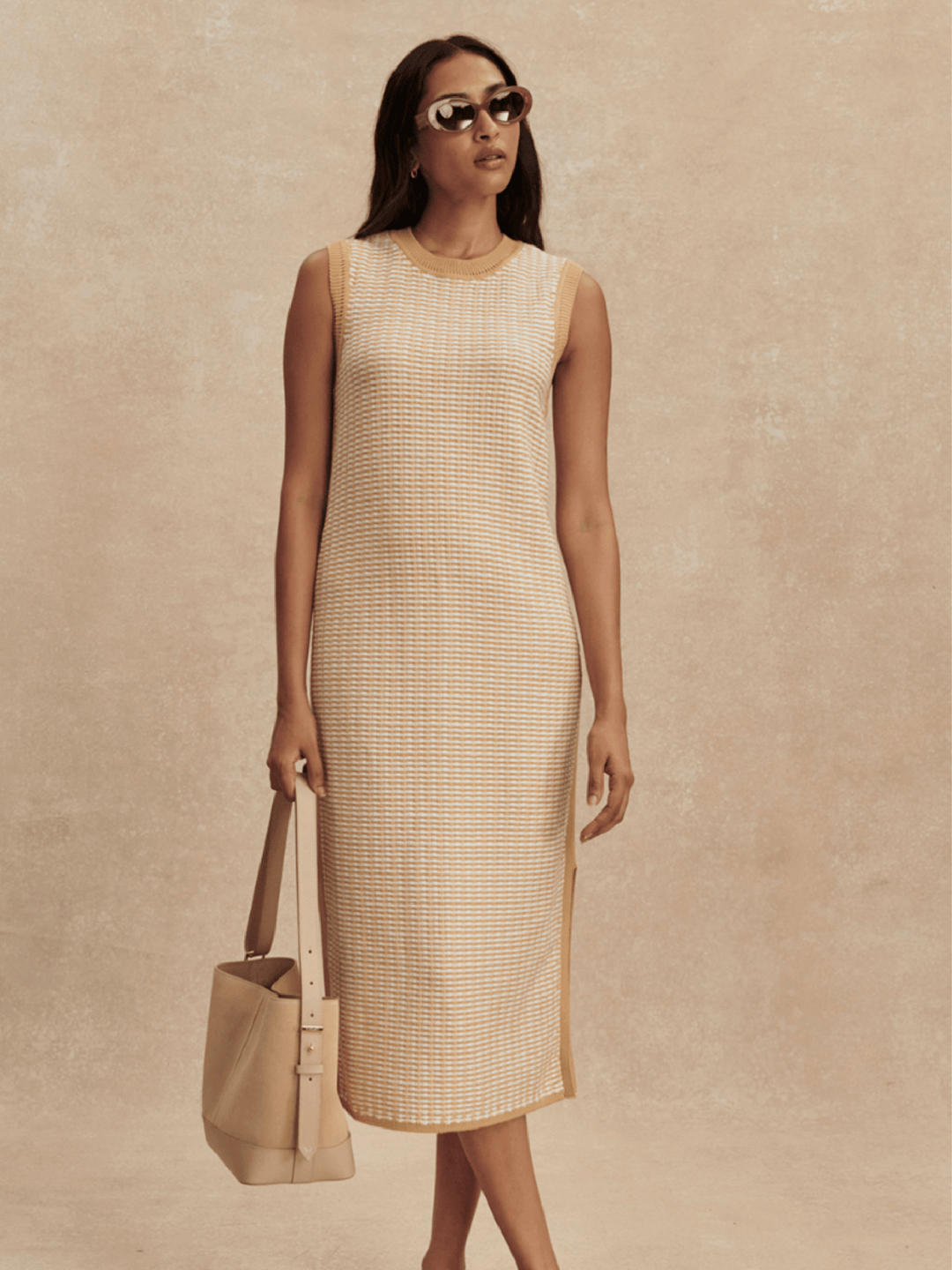 Woman wearing a beige sleeveless dress holding a matching handbag against a beige background