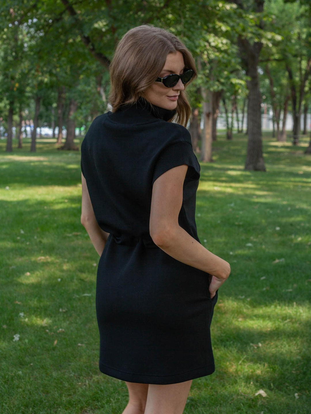 A woman wearing a black sleeveless turtleneck dress with side pockets, standing in a park with trees and grass in the background.
