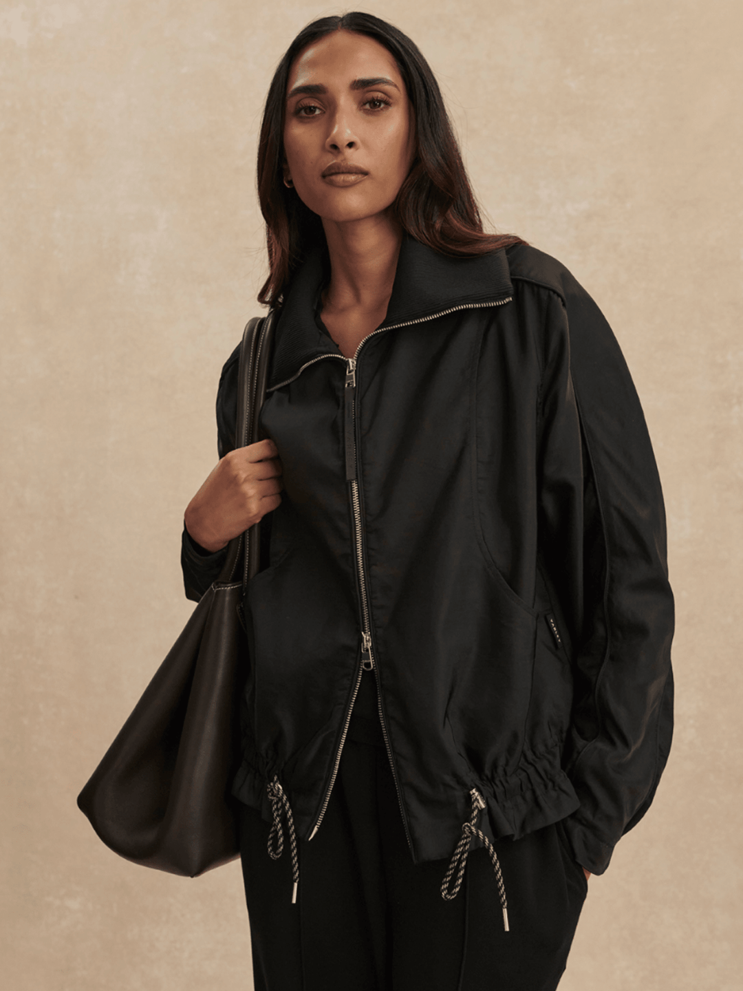 Woman wearing a black jacket and holding a matching bag against a beige background