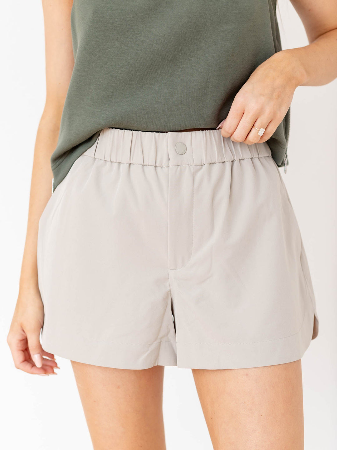 Woman wearing a green sleeveless top and beige shorts against a white wall.
