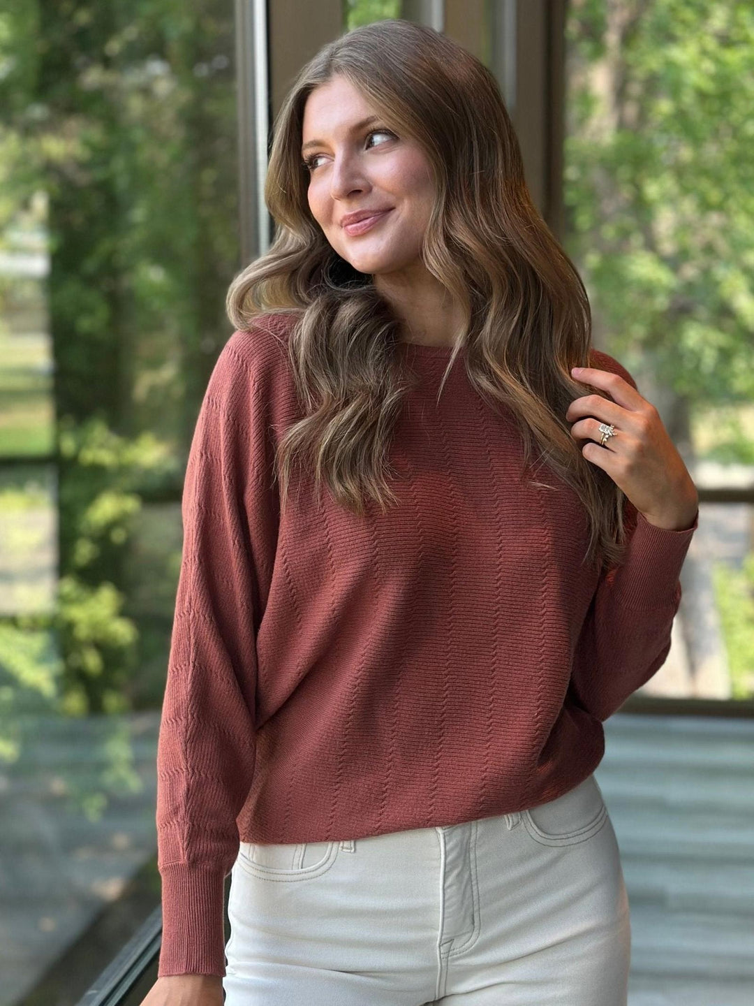 A woman wearing a textured, dusty rose long-sleeve knit sweater and white pants, standing in front of a window with a blurred green outdoor background.