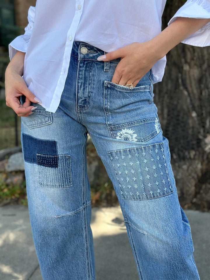 A person wearing light blue patchwork denim jeans with multiple pockets, including a star-patterned patch and a dark blue patch on the thigh, paired with a white button-down shirt in an outdoor setting.