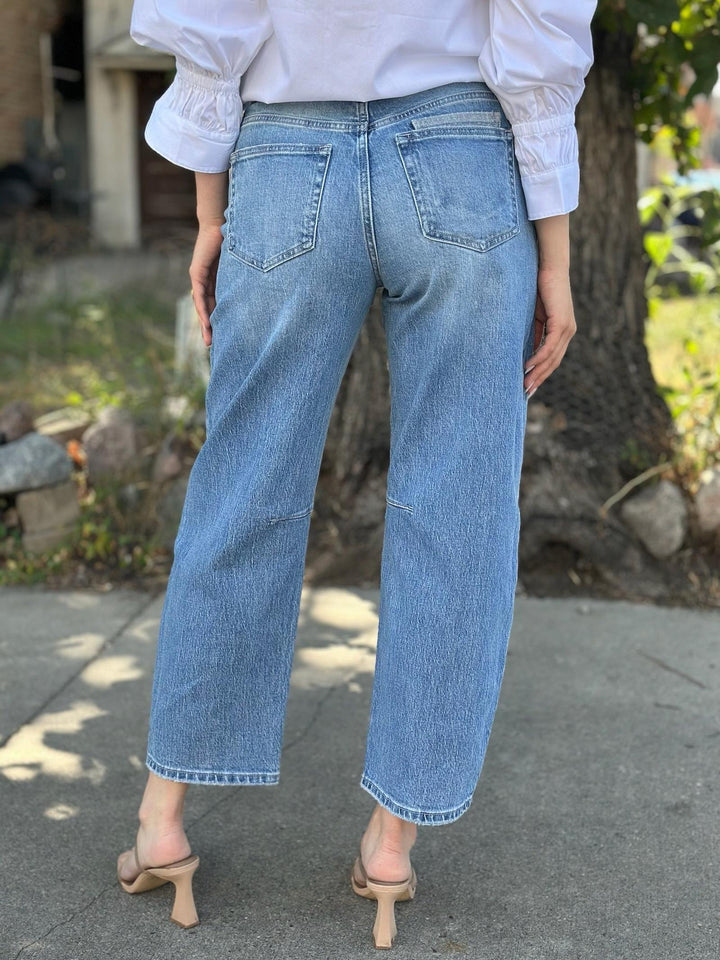 A person shown from the back wearing light-wash blue wide-leg cropped jeans, a white blouse, and beige heeled sandals, standing outdoors near a tree and pavement.