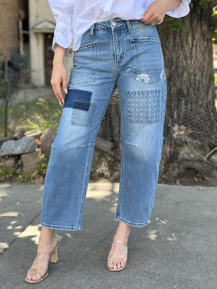 A person wearing light-wash blue patchwork denim jeans with dark blue contrast patches and distressed details, styled with a white blouse and clear-strap heeled sandals outdoors.