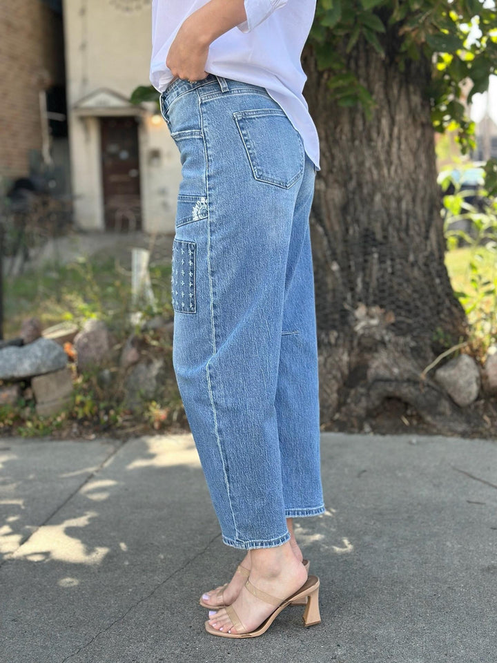 A person wearing light-wash blue cropped jeans with a distressed patch detail on the left thigh, paired with a white top and beige heeled sandals, standing outdoors near a tree.