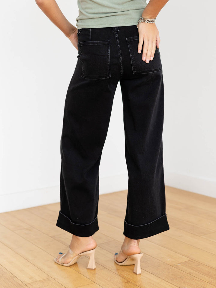 Person wearing black wide-leg jeans on a wooden floor with a white wall background