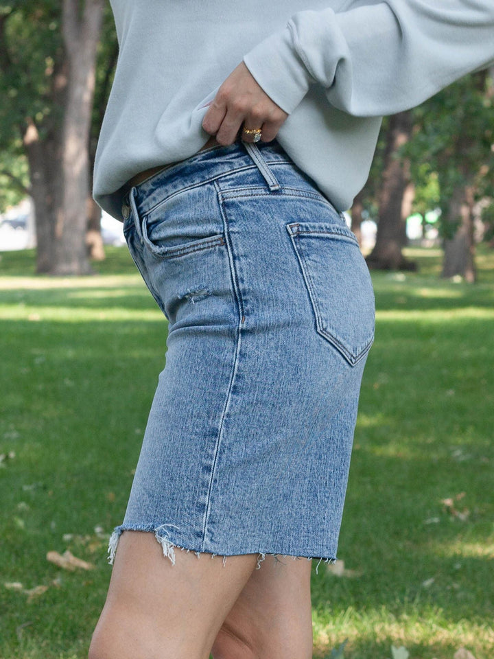 A person outdoors in a grassy park wearing a light-wash blue denim mini skirt with a raw hem and a light gray sweatshirt.