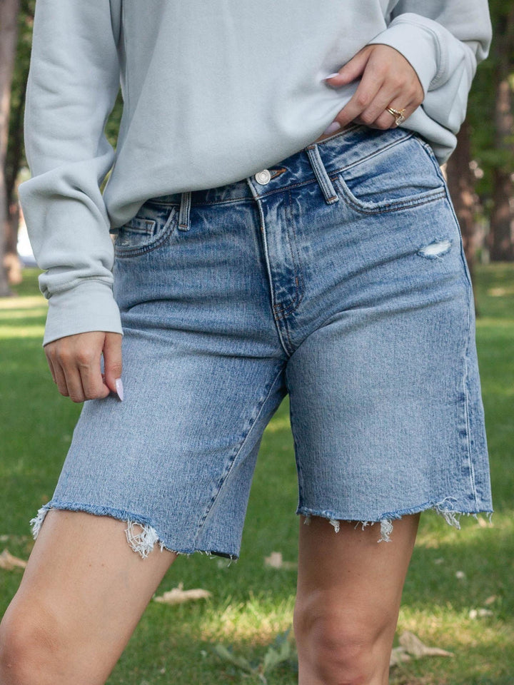 A person outdoors wearing light-wash blue denim shorts with a raw frayed hem and a light gray sweatshirt, standing on grass.