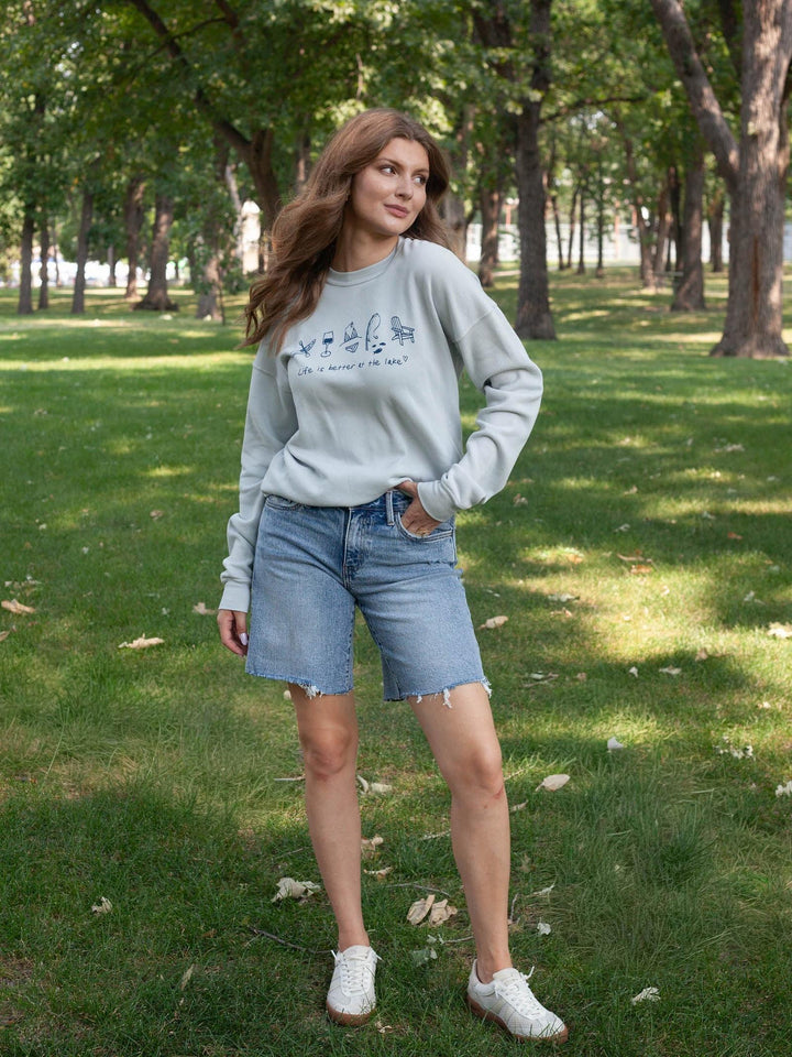 A woman stands in a park wearing a light gray sweatshirt with a dark graphic print, light-wash denim shorts, and white sneakers.