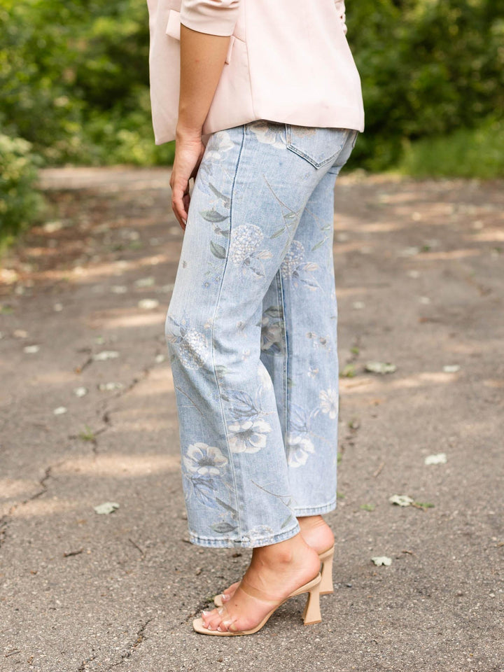 A person outdoors wearing light-wash blue cropped jeans with a faded floral pattern, paired with a pale pink top and clear-strap heeled sandals on a paved path with green foliage in the background.