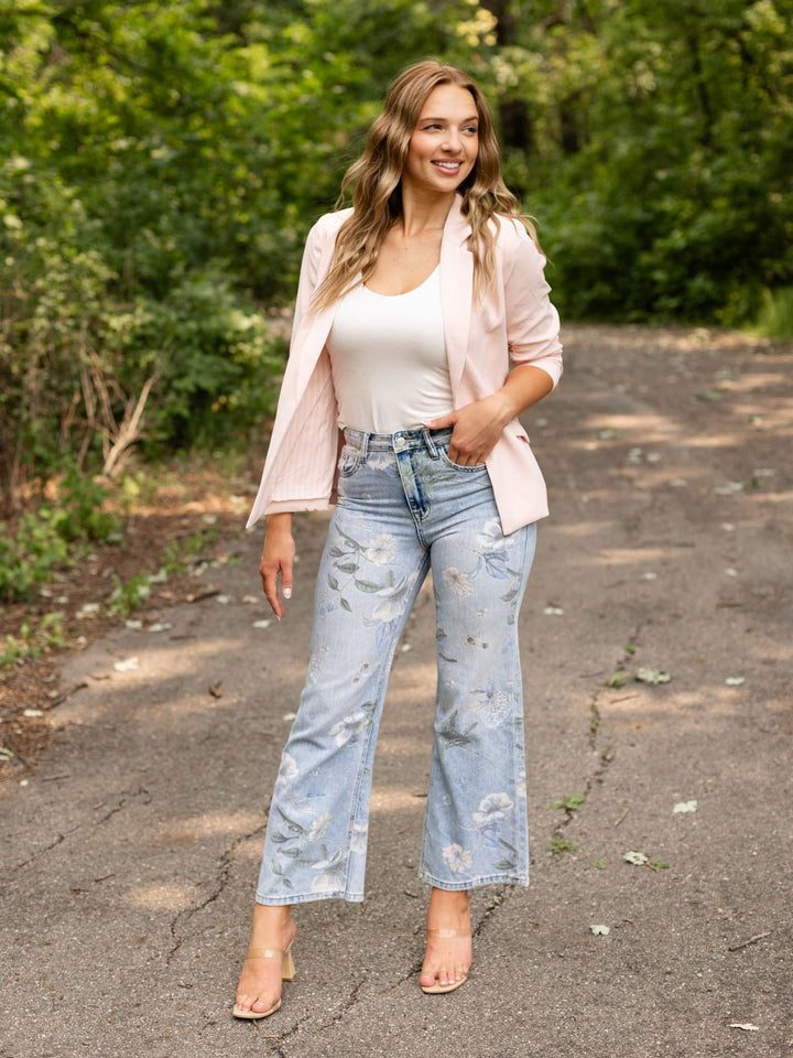 A woman stands on an outdoor paved path surrounded by green trees, wearing a light pink open-front blazer over a white tank top, light-wash distressed flare jeans, and clear high-heel sandals.