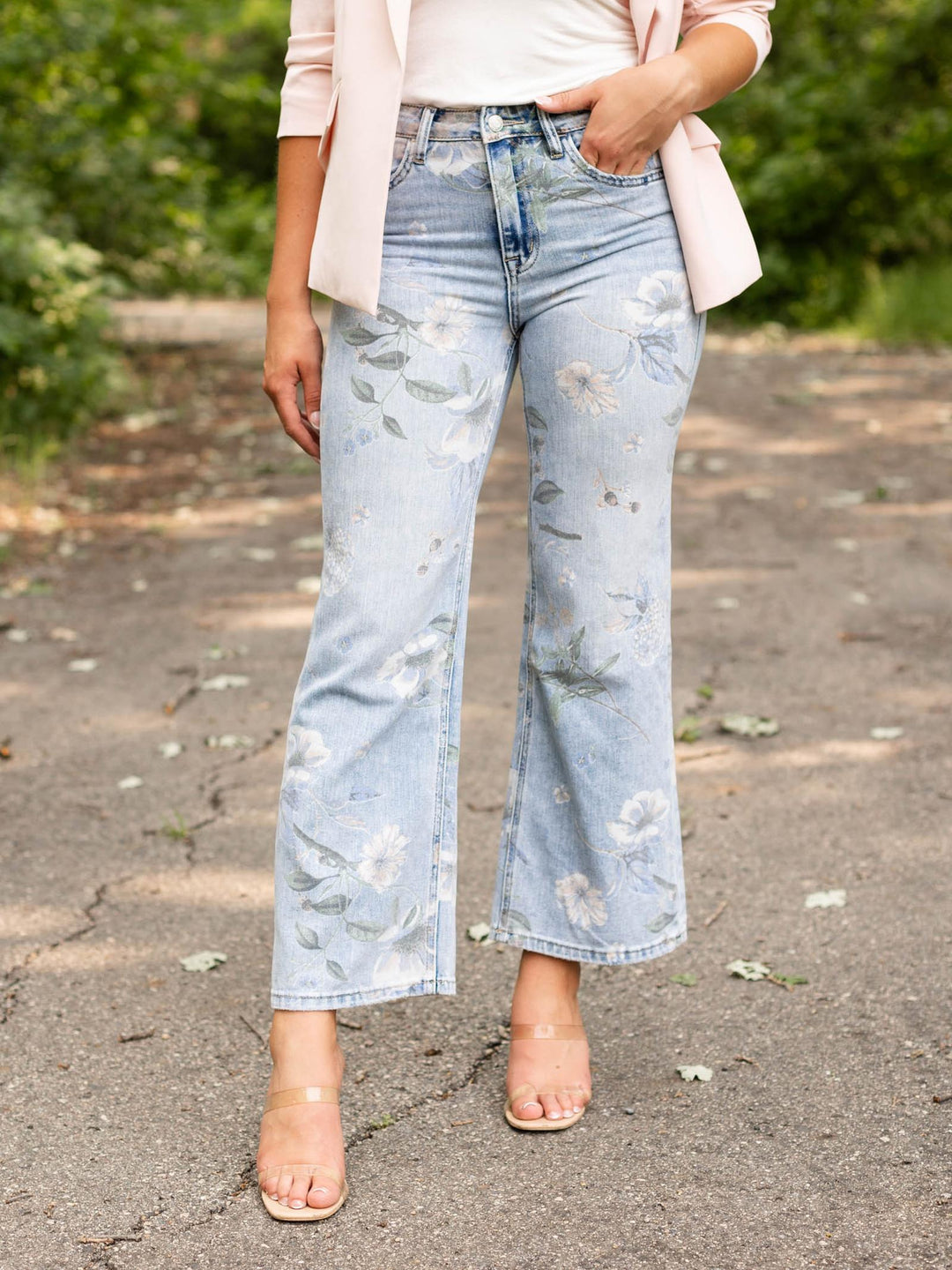 A person outdoors wearing light-wash blue cropped flare jeans with a faded floral print, paired with a pale pink blazer and clear high-heel sandals.