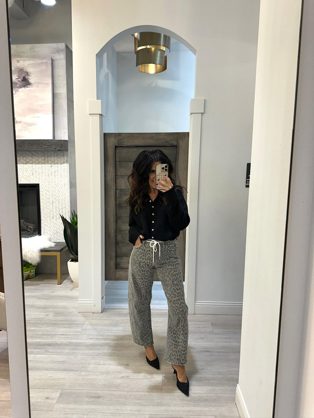 Person taking a mirror selfie wearing a black shirt and gray patterned pants in a room with white walls and wooden flooring.
