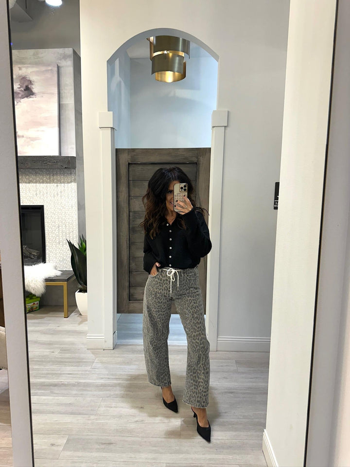 Person taking a mirror selfie wearing a black shirt and gray patterned pants in a room with white walls and wooden flooring.