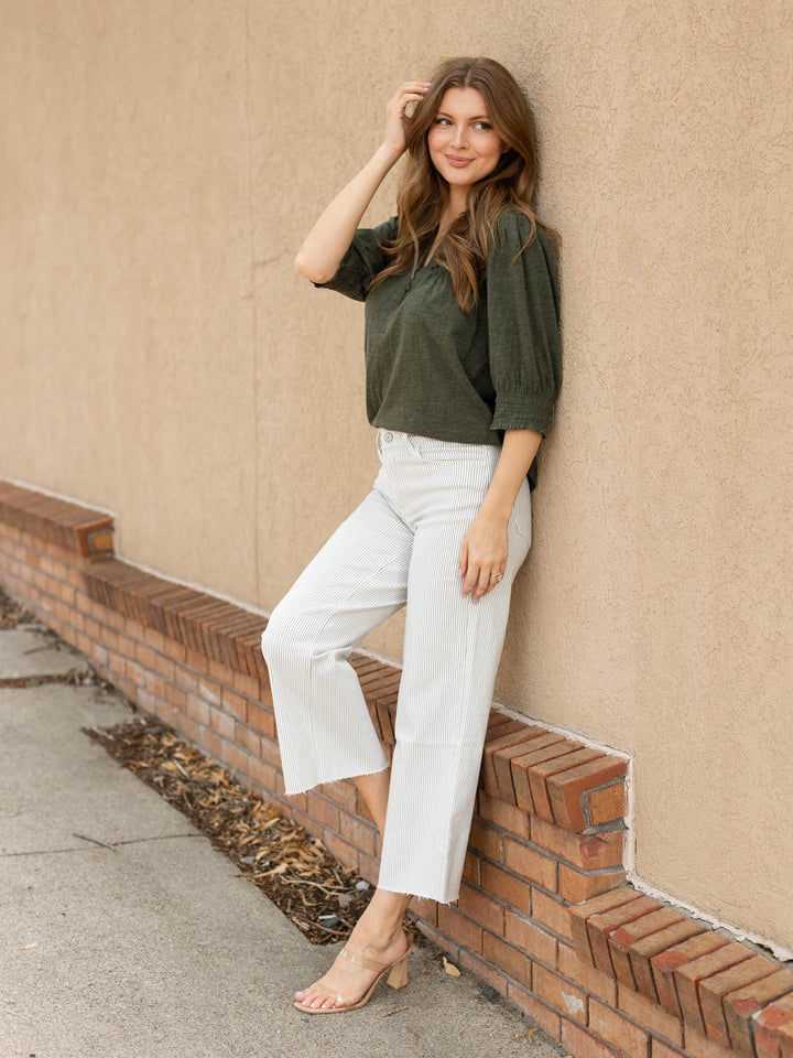 A woman posing outdoors against a beige stucco wall and brick base, wearing an olive green short-sleeve top, white cropped pants, and clear high-heeled sandals.