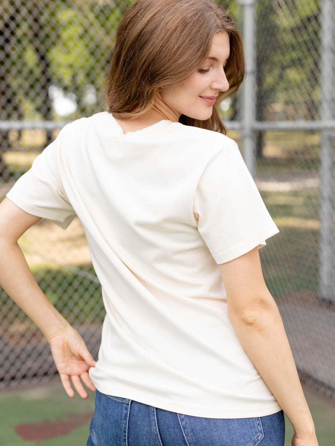 A woman wearing a cream-colored short-sleeve t-shirt and blue jeans, photographed from behind in an outdoor setting with a chain-link fence in the background.