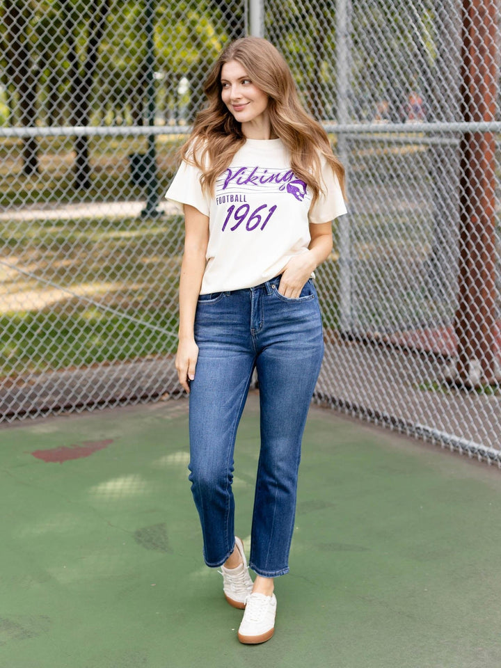 A woman stands on an outdoor court in front of a chain-link fence, wearing a cream graphic T-shirt with purple text reading &
