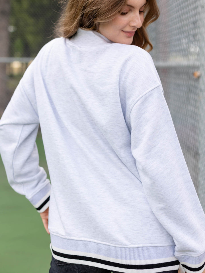 A woman wearing a light heather gray sweatshirt with black and white striped ribbed cuffs and hem, photographed outdoors on a tennis court.