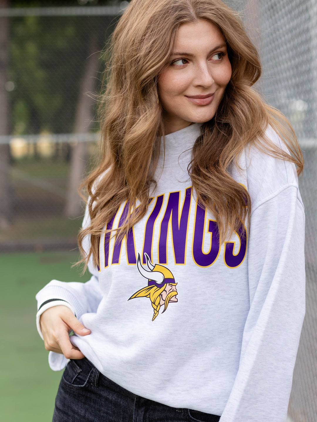 A woman wearing a white Minnesota Vikings sweatshirt with purple and gold lettering, standing outdoors in front of a chain-link fence.
