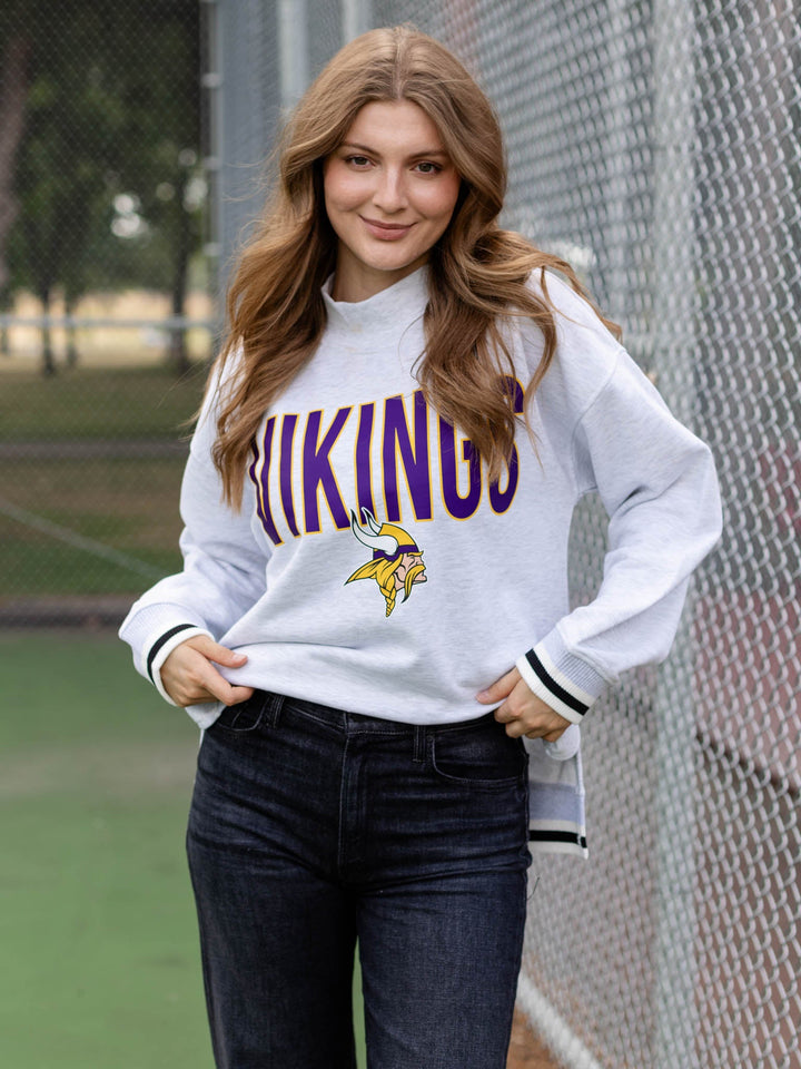 A woman wearing a white Minnesota Vikings sweatshirt with purple and yellow lettering, paired with dark blue jeans, standing in front of a chain-link fence.