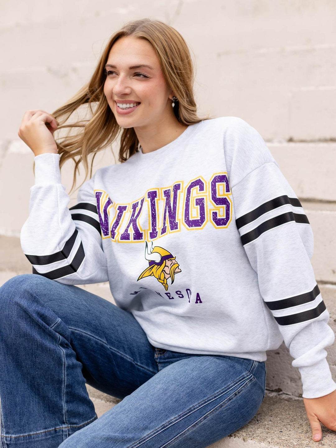 A woman wearing a white Minnesota Vikings sweatshirt with purple and yellow lettering and black and white striped sleeves, paired with blue jeans, sitting against a light-colored brick wall.