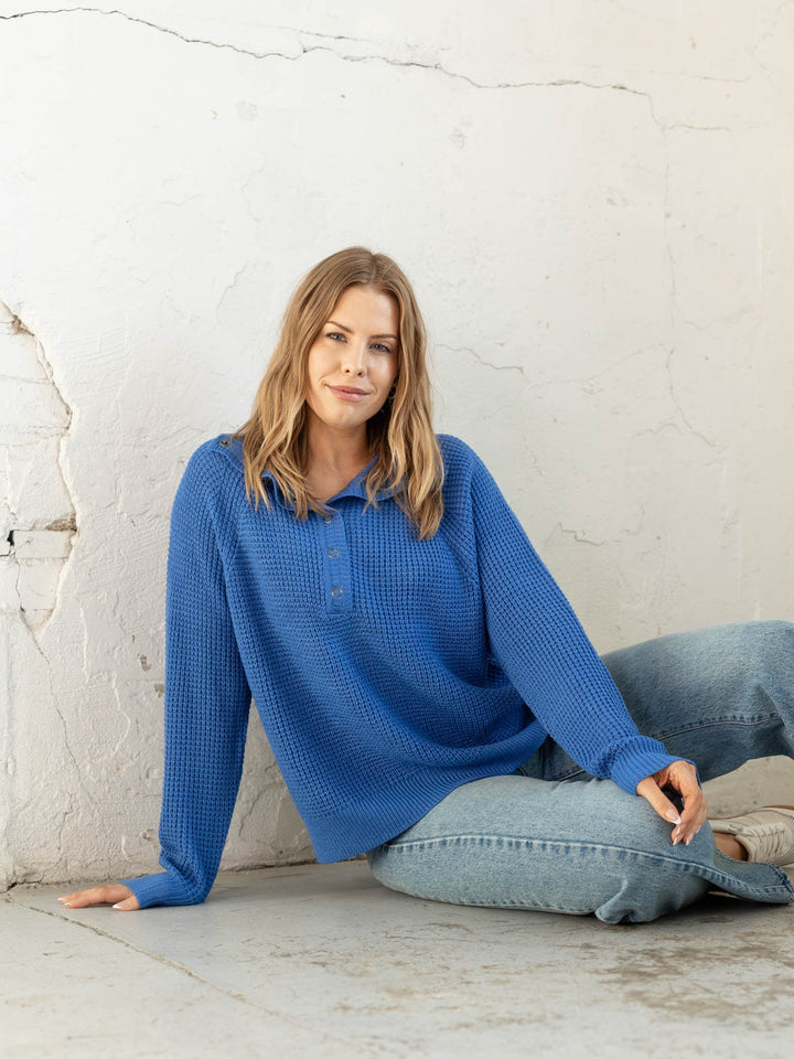 Woman wearing a blue sweater sitting against a white brick wall.