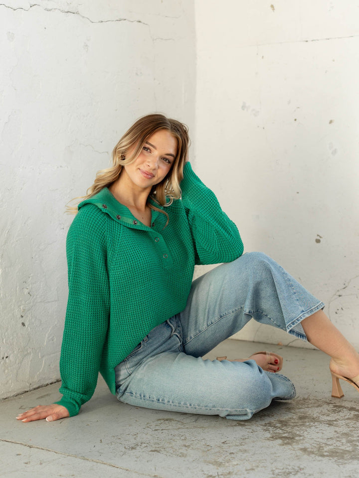 Woman wearing a green sweater and blue jeans sitting against a white wall.