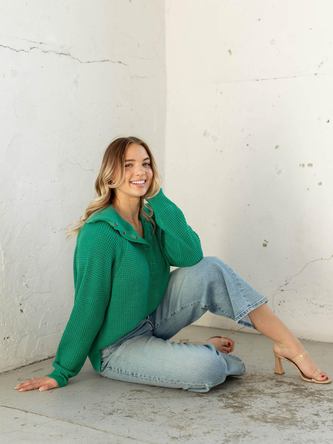 Woman in a green sweater and jeans sitting against a white wall.