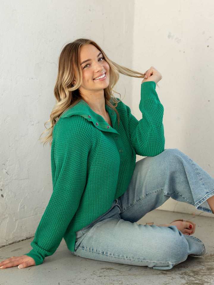 Woman wearing a green sweater and blue jeans sitting on the floor against a white wall.