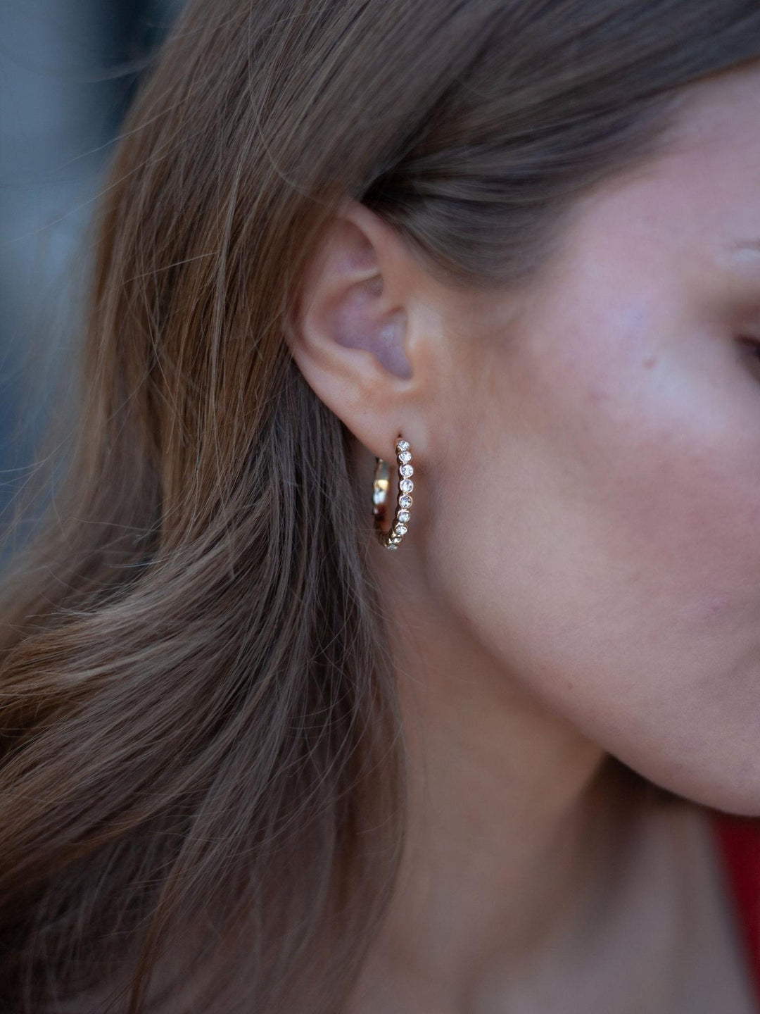 A close-up side profile of a woman wearing a small gold huggie hoop earring adorned with a row of clear pavé-set stones.