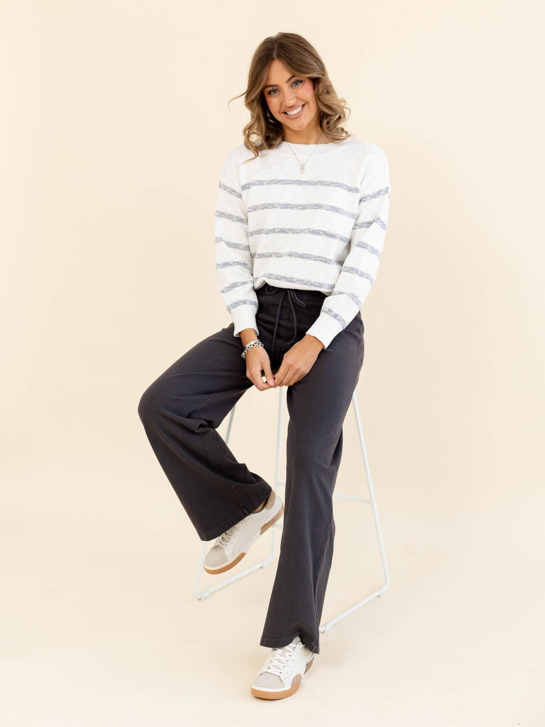 Woman wearing a striped sweater and dark pants sitting on a chair against a beige background