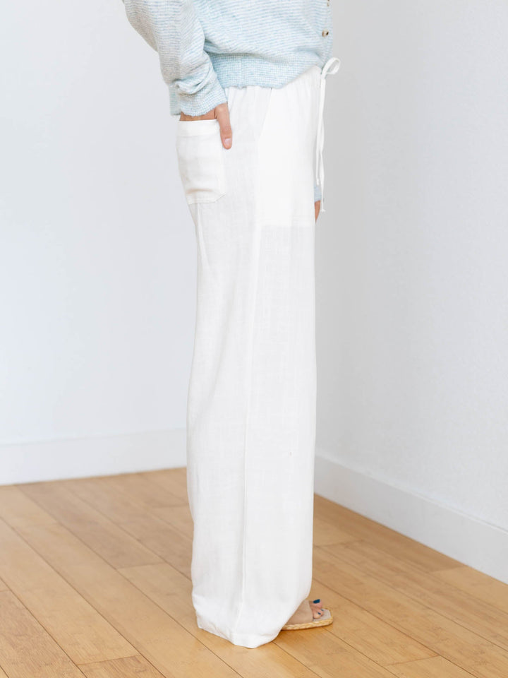 Person wearing light blue sweater and white pants standing on a wooden floor with a white wall background