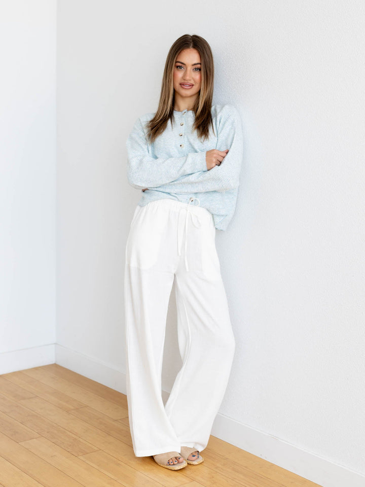 Woman wearing a light blue sweater and white pants standing against a white wall.
