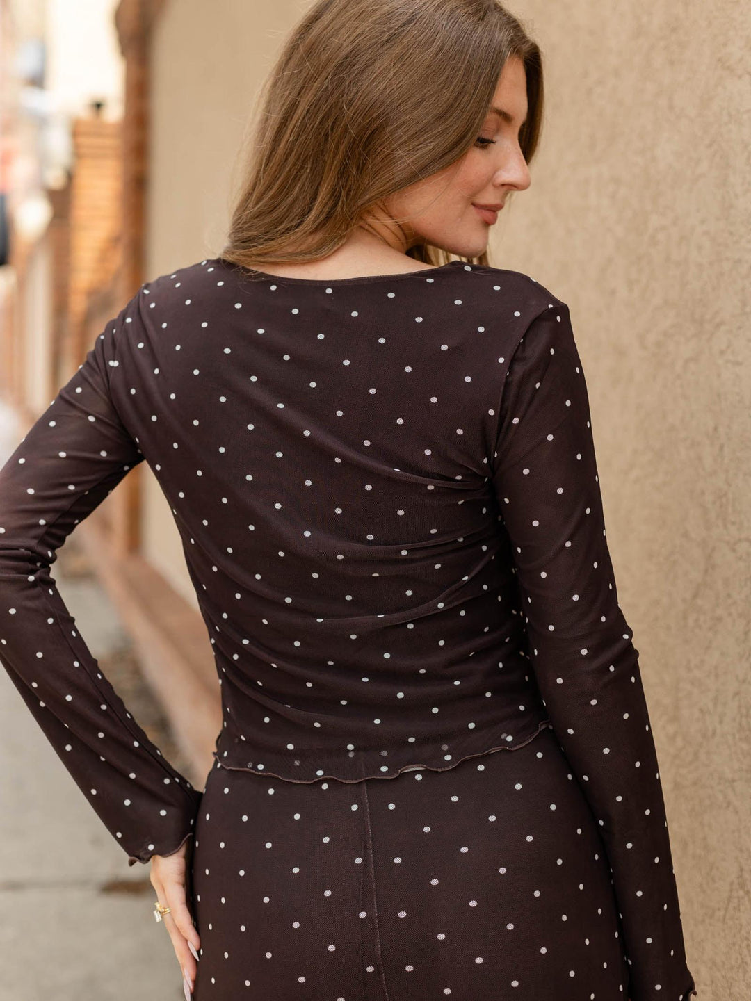 A woman wearing a dark brown long-sleeve dress with a white polka dot pattern, shown from a rear three-quarter view against a beige stucco wall.