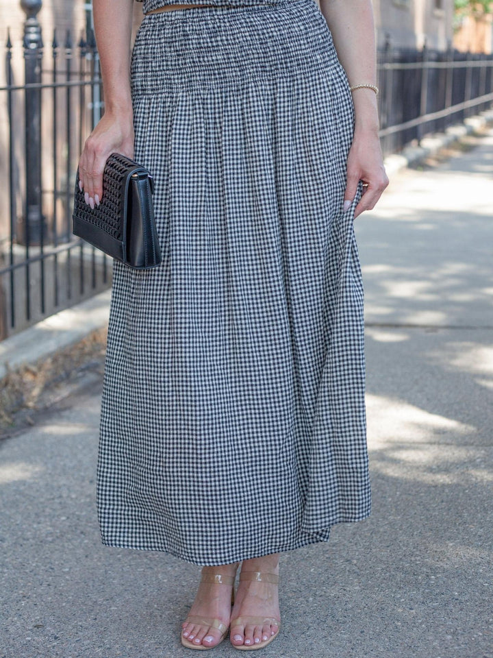 A person standing outdoors on a sidewalk wearing a black and white gingham pleated maxi skirt, holding a black clutch, and wearing clear high-heel sandals.