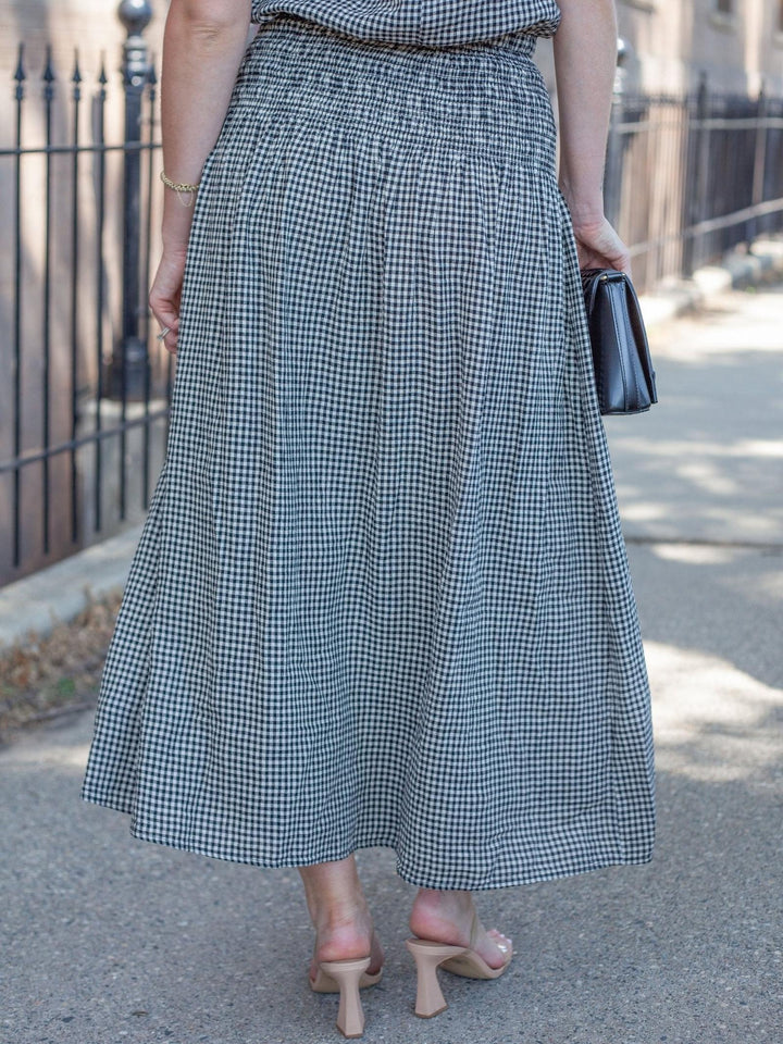 A person outdoors wearing a black and white gingham pleated maxi skirt, paired with beige heeled sandals and holding a small black clutch.