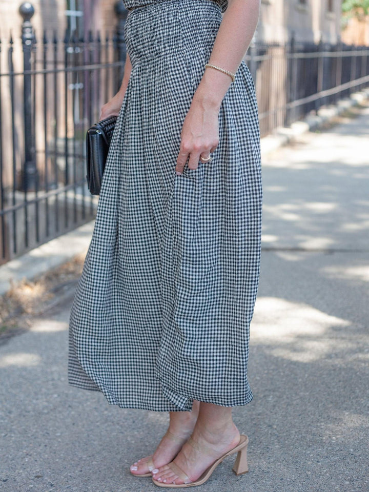A person standing outdoors on a sidewalk wearing a black and white gingham midi skirt, holding a black clutch, and wearing beige block-heel mule sandals.