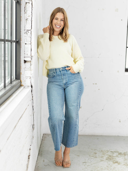 Woman wearing a light-colored sweater and blue jeans standing against a white wall.