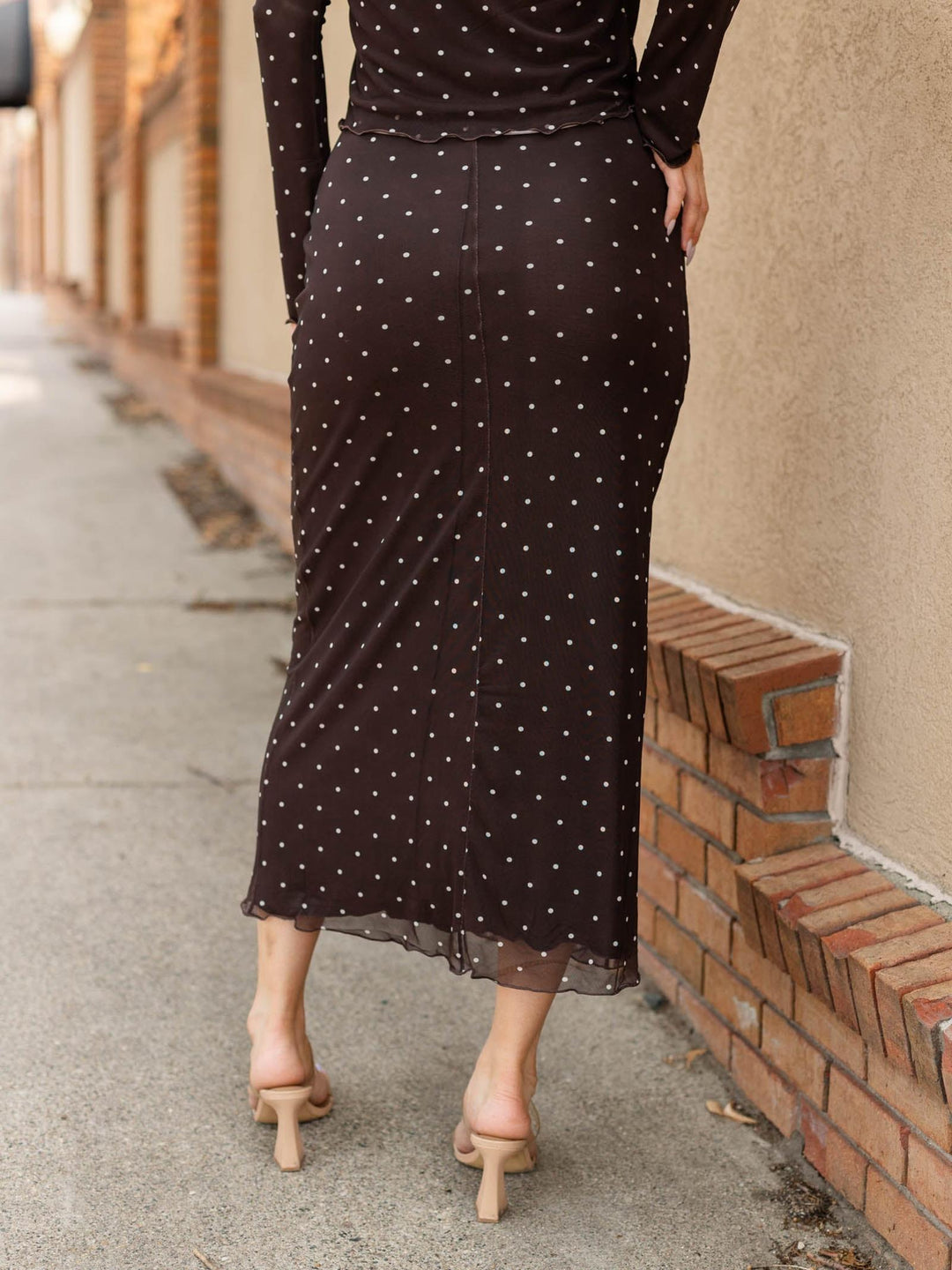 A woman wearing a dark brown long-sleeve midi skirt with white polka dots, shown from behind as she walks along a sidewalk next to a brick wall.