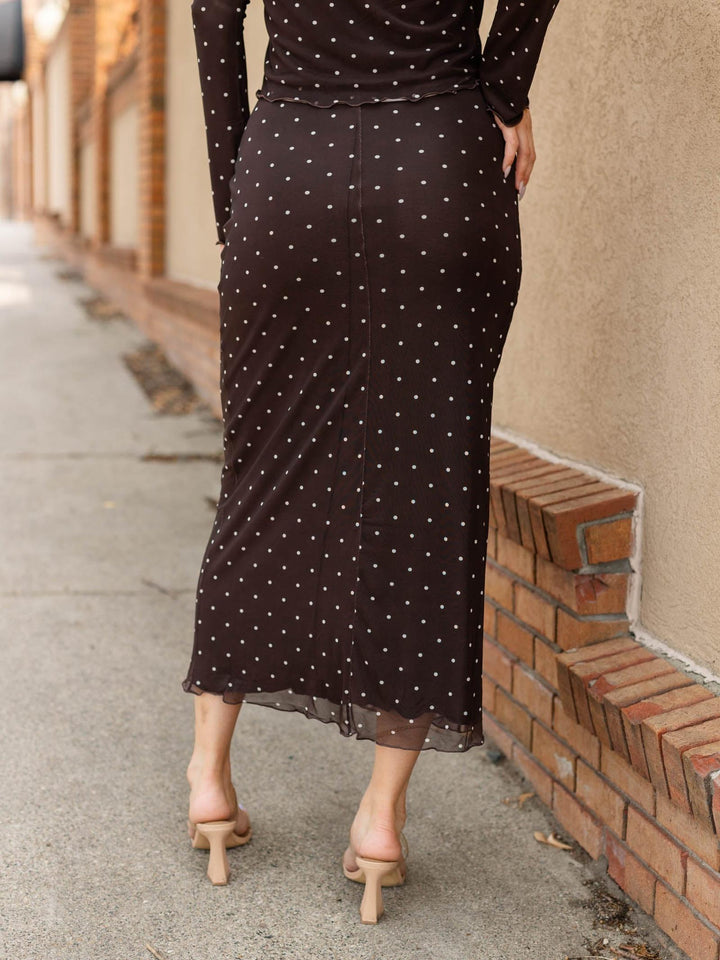 A woman wearing a dark brown long-sleeve midi skirt with white polka dots, shown from behind as she walks along a sidewalk next to a brick wall.