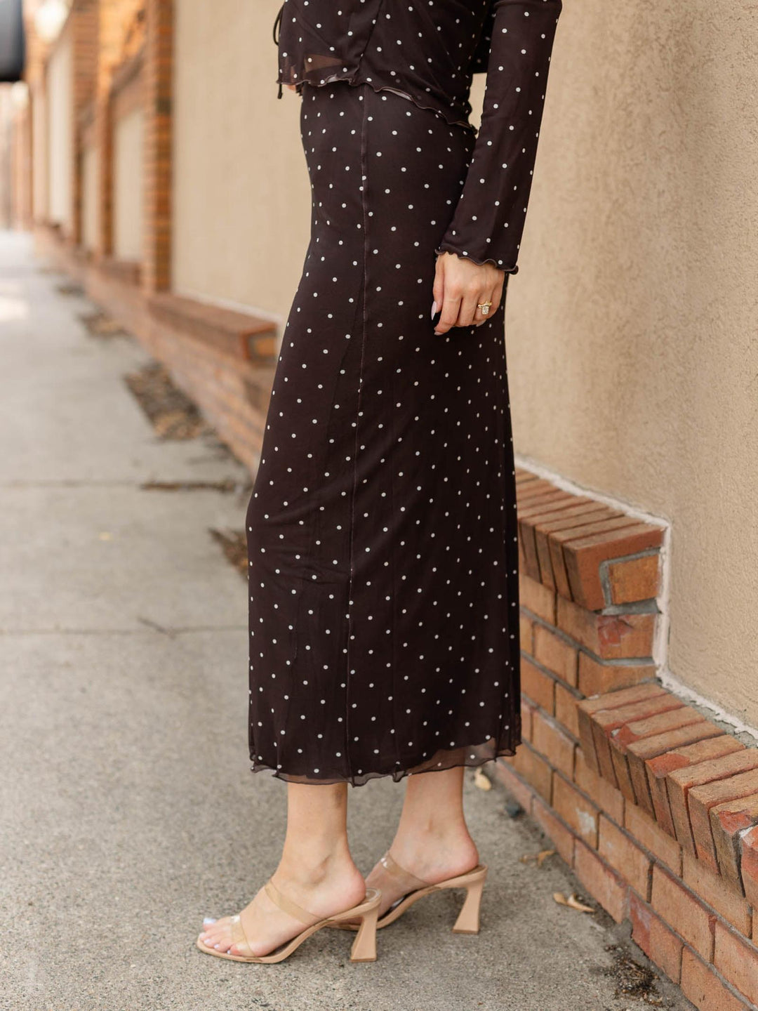 A woman wearing a black midi skirt with white polka dots and a matching long-sleeve top, paired with clear strap heeled sandals, standing on a sidewalk next to a brick wall.